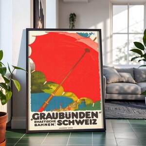 Puede incluir: Póster de viaje vintage enmarcado con un gran paraguas rojo sobre un fondo de colinas verdes y agua azul. El póster tiene texto en alemán: "GRAUBÜNDEN SCHWEIZ". El póster está en un marco negro y se apoya contra una pared.