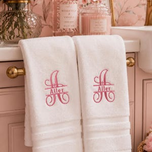 May include: Two white hand towels hanging on a gold towel bar, each featuring a pink embroidered monogram of the letter A with the name Aller stitched across the center. The towels have a textured border detail at the bottom edge.