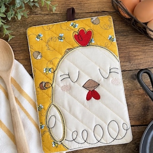 Puede incluir: Un agarrador de cocina acolchado rectangular con fondo amarillo y un patrón de pequeñas abejas y bellotas. El centro muestra una cara de pollo blanca con ojos cerrados, pico marrón, cresta roja y barbilla roja, con una presilla para colgar.