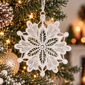 May include: A white lace snowflake ornament featuring intricate threadwork and a single clear crystal at the center. The six-pointed star design is suspended by a thin white ribbon loop, showcasing detailed embroidery patterns throughout the structure.