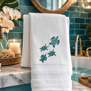 May include: A white hand towel featuring an embroidered pattern of four sea turtles in a teal blue color. The towel has a soft, plush texture with a simple hemmed edge and three horizontal decorative lines stitched near the bottom border.
