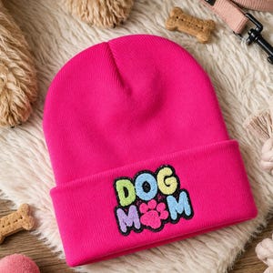 May include: A bright pink knit beanie featuring an embroidered patch on the cuff with the text DOG MOM in colorful letters and a central pink paw print. The beanie is made of a ribbed fabric and has a folded brim design.