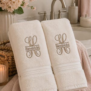 May include: Two folded cream-colored bath towels featuring an embroidered monogram design in a light brown thread. The embroidery includes the name Hendricks centered between two stylized, symmetrical scrollwork patterns on each towel.