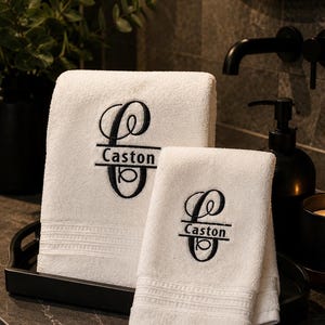 以下が含まれることがあります： 中央に「Caston」という名前が水平線で刺繍された、黒いモノグラム付きの白いコットンタオル2枚。タオルは下部に質感のあるボーダーのディテールが施されており、バスルームの黒いトレイの上に折りたたんで置かれています。