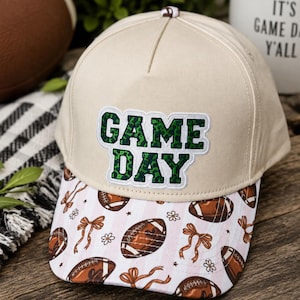 May include: A beige baseball cap featuring a white patch with the green leopard-print text GAME DAY. The brim displays a repeating pattern of brown footballs, orange bows, and small white flowers against a background of light pink stripes.
