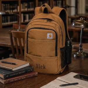 May include: A tan Carhartt backpack featuring a front zippered pocket, a black mesh side pocket, and the name Nick embroidered in dark brown thread on the lower front section. The bag has a top handle, adjustable shoulder straps, and black zippers.