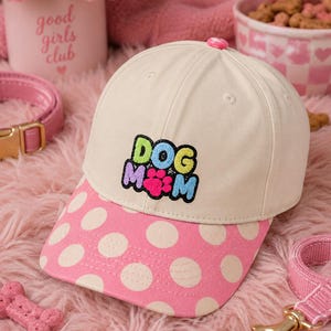May include: A cream-coloured baseball cap with a pink polka-dot brim. The front features an embroidered patch with the text DOG MOM in colourful letters and a bright pink paw print in the centre. The cap has a small pink button on the top crown.