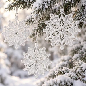 May include: Three white lace snowflake ornaments hanging from a pine tree. Each ornament features intricate threadwork patterns and a small, clear, circular crystal bead at the center. The snowflakes are suspended by thin white ribbons.