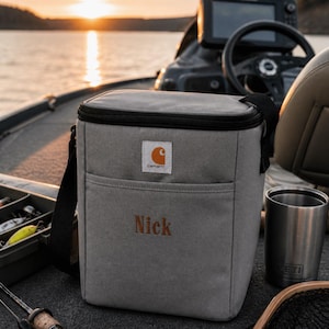 May include: A gray Carhartt cooler bag with a black shoulder strap and the name "Nick" embroidered in orange. The bag has a front pocket and a black zipper. A stainless steel tumbler and fishing gear are also visible.
