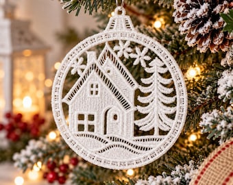 May include: A white circular lace ornament featuring an intricate embroidered design of a small house with a pitched roof, a tall evergreen tree, and several small snowflakes, all enclosed within a decorative circular border with a metal loop.