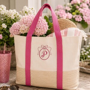 Pode incluir: Uma bolsa tote de lona na cor creme com alças de tecido rosa vibrante e uma base correspondente com textura trançada. A parte frontal da bolsa exibe um emblema circular rosa bordado com um detalhe de laço e a letra P no centro.