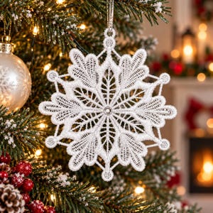May include: A white snowflake ornament crafted from intricate lace embroidery. The design features a symmetrical, star-like pattern with delicate, open-work stitching and a small, clear circular bead centered at the core, suspended by a silver cord.