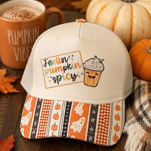 Op de afbeelding: Een crèmekleurige truckerpet met een geborduurde rechthoekige patch met de tekst "Feelin' pumpkin spicy" en een cartoon koffiebeker. De klep heeft een herhalend patroon van witte spoken, oranje pompoenen en geruite strepen.