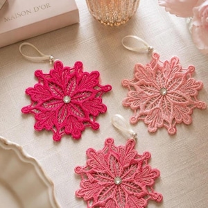 May include: Three snowflake-shaped ornaments crafted from intricate pink, fuchsia, and light pink lace. Each ornament features a central clear rhinestone and a white ribbon loop for hanging, showcasing detailed embroidery and delicate floral patterns.