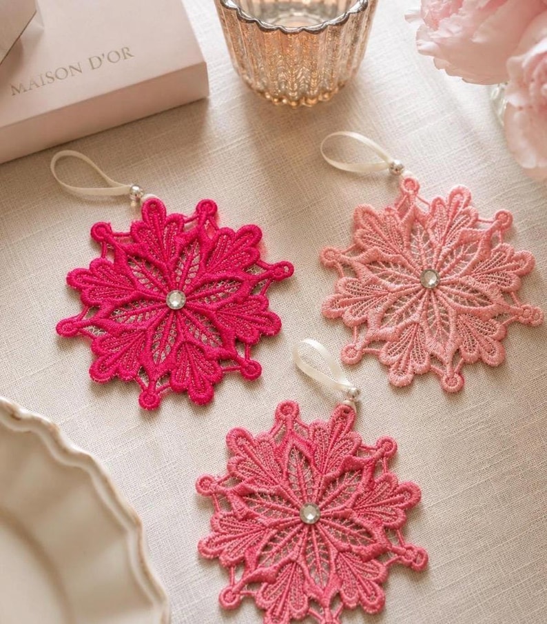 May include: Three snowflake-shaped ornaments crafted from intricate pink, fuchsia, and light pink lace. Each ornament features a central clear rhinestone and a white ribbon loop for hanging, showcasing detailed embroidery and delicate floral patterns.