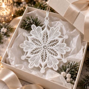 May include: A white lace snowflake ornament featuring intricate threadwork and a single clear crystal at the center. The ornament is suspended by a thin white ribbon and rests inside a square gift box lined with white tissue paper.