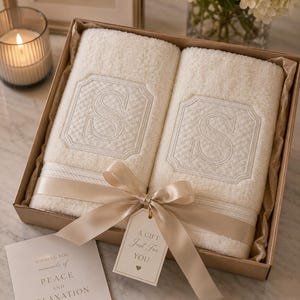 Puede incluir: Dos toallas de rizo color crema con una letra S bordada dentro de un borde decorativo, presentadas en una caja de regalo de cartón atada con una cinta de satén beige y una etiqueta que dice A GIFT Just For YOU, junto a una tarjeta con texto.