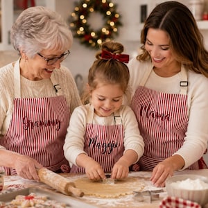 May include: Three matching red and white striped aprons featuring embroidered text. The left apron displays the word Grammy, the center apron displays the name Maggie, and the right apron displays the word Mama. Each apron has white straps.