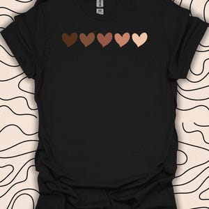 May include: Black t-shirt featuring a row of six hearts in different shades of brown across the chest. The hearts represent different skin tones.