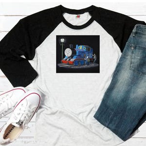 Könnte beinhalten: Weißes und schwarzes Raglan-Baseball-T-Shirt mit einem Cartoon-Zug-Motiv. Der blaue Zug hat ein freundliches Gesicht. Das T-Shirt wird mit blauen Jeans und weißen Turnschuhen kombiniert.