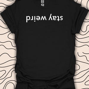 Może przedstawiać: Czarny T-shirt z białym tekstem, który brzmi "Stay Weird" w lustrzanej czcionce.