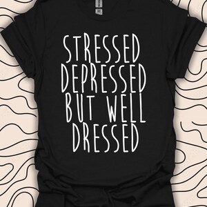 Op de afbeelding: Zwart T-shirt met witte tekst die "Stressed Depressed But Well Dressed" luidt.