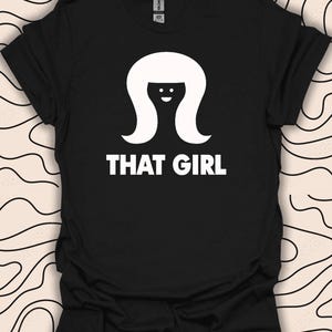 May include: Black t-shirt featuring a white graphic of a woman with a long, straight hairstyle and the text "THAT GIRL".