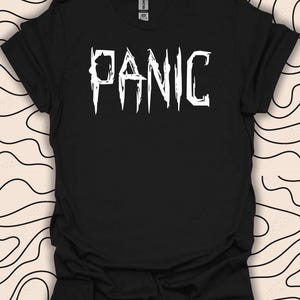 Może przedstawiać: Czarny T-shirt z nadrukiem słowa "PANIC" w kolorze białym, w stylu z ząbkowanymi krawędziami.