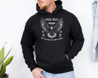 May include: Black hooded sweatshirt with a white graphic of an eagle with outstretched wings. The text "FREE BIRD" is above the eagle, with "1973" and the phrase "AND THIS BIRD YOU CANNOT CHANGE" below. The sweatshirt has a front pocket and a drawstring hood.