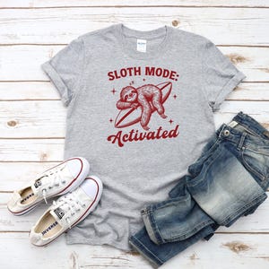 Może przedstawiać: Szara koszulka z napisem "Sloth Mode: Activated" i czerwoną grafiką leniwca na desce surfingowej. Koszulka jest połączona z niebieskimi dżinsami i białymi trampkami.