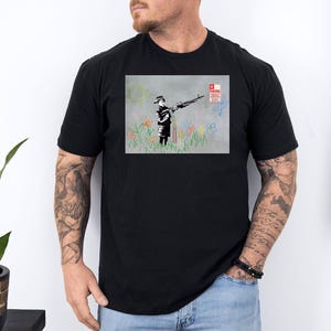 Könnte beinhalten: Schwarzes T-Shirt mit einem Druck eines Banksy-Kunstwerks. Das Kunstwerk zeigt eine Figur, die eine Waffe hält, die Blumen sprüht. Der Hintergrund umfasst bunte Blumen, eine Sonne und ein "No Parking"-Schild.