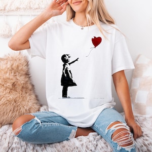 Könnte beinhalten: Weißes T-Shirt mit einer schwarzen Silhouette eines Kindes, das nach einem roten herzförmigen Ballon greift. Das Design erinnert an den Stil von Banksy. Das Shirt hat eine lockere, übergroße Passform.