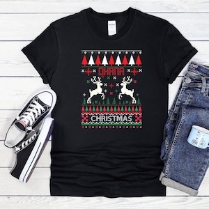 以下が含まれることがあります： 黒いTシャツに、お祝いのクリスマスのデザイン。赤いと白の木、トナカイ、そして「OHANA CHRISTMAS」の言葉がピクセル化されたスタイルで描かれています。黒と白のスニーカーとブルージーンズがシャツの隣にあります。
