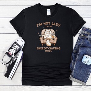 Może przedstawiać: Czarny t-shirt z kreskówkowym kotem w fotelu, trzymającym pilota i napój. Tekst brzmi: "I'M NOT LAZY I'M IN ENERGY-SAVING MODE." Zawiera również czarno-białe trampki i niebieskie dżinsy.