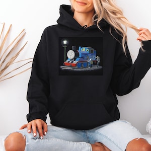 Puede incluir: Sudadera con capucha negra con una colorida ilustración de un tren de dibujos animados azul con una cara sonriente, una farola y dos pequeñas figuras. El tren tiene detalles en rojo y azul. La sudadera tiene un bolsillo delantero y una capucha con cordón.