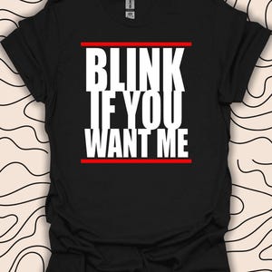Op de afbeelding: Zwart T-shirt met witte tekst die "Blink if you want me" luidt met een rode lijn boven en onder de tekst.