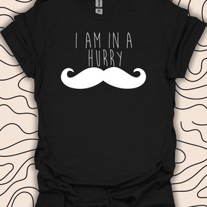 Op de afbeelding: Zwart T-shirt met de witte tekst "I AM IN A HURRY" boven een grote witte snorafbeelding. Het T-shirt heeft korte mouwen en een ronde hals. De achtergrond is crèmekleurig met een zwart golvend patroon.