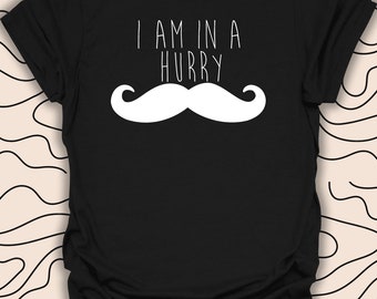 May include: Black t-shirt with the white text "I AM IN A HURRY" above a large white mustache graphic. The shirt is a short-sleeved crew neck style. The background has a cream color with a black wavy pattern.