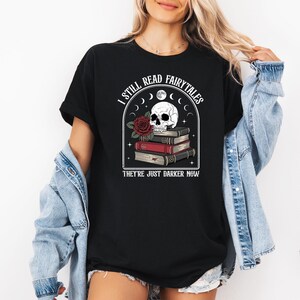 Puede incluir: Camiseta negra con un gráfico que presenta una calavera sobre una pila de libros, una rosa roja y el texto "I STILL READ FAIRYTALES THEY'RE JUST DARKER NOW." La camiseta se usa con una chaqueta vaquera azul.