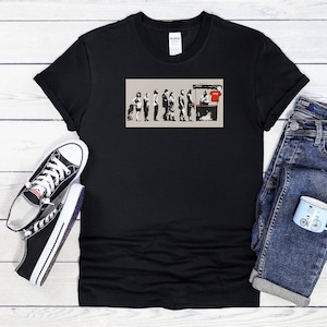 Puede incluir: Camiseta negra con un gráfico de personas haciendo cola en un puesto, con una persona con una camisa roja. La imagen se combina con vaqueros azules, zapatillas blancas y negras y una pequeña taza azul.