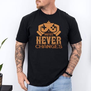 May include: Black t-shirt with a brown graphic of a video game controller and the text "WAR NEVER CHANGES". The shirt is made of a soft material and has short sleeves.