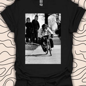 Peut inclure: Une photographie en noir et blanc d'un chimpanzé à vélo. Le chimpanzé porte un maillot de course blanc et noir avec le numéro 22. Le chimpanzé roule sur un trottoir avec deux enfants qui marchent derrière lui.