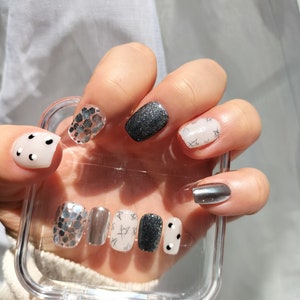 Könnte beinhalten: Ein durchsichtiger Plastikbehälter mit verschiedenen Designs für künstliche Nägel. Die Designs umfassen schwarzen Glitzer, silbernen Glitzer, Weiß mit schwarzen Sternen und Weiß mit schwarzen Punkten.