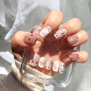 Könnte beinhalten: Nahaufnahme einer Hand mit nude-farbenen Press-on-Nägeln, die mit kleinen roten Kirschen und weißen Punkten verziert sind. Die Nägel werden über ein transparentes Handy-Case gehalten.