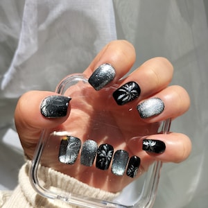 Könnte beinhalten: Ein Set mit schwarzen und silbernen Press-on-Nägeln mit Palmenmotiv. Die Nägel werden in einem durchsichtigen Plastikbehälter gezeigt.