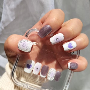 Könnte beinhalten: Lila und weißer Nagellack mit Herz- und Glitzerdesigns.