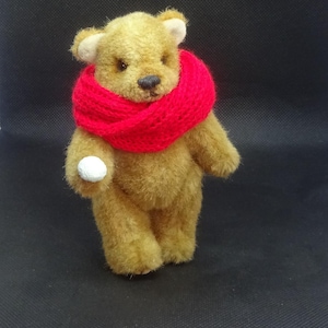 May include: A soft toy teddy bear with tan fur and a vibrant red scarf. The bear is holding a small white ball. The bear has a black nose and small, round ears. The background is a solid black.