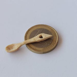 Puede incluir: Una pequeña cuchara de madera hecha a mano con un acabado natural. La cuchara presenta un cuenco tallado y un mango con un agujero decorativo. La cuchara está junto a una moneda como referencia de tamaño.