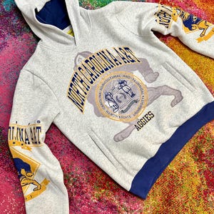 May include: A light gray hooded sweatshirt featuring North Carolina A&T branding. The front displays the university seal and the text North Carolina A&T Aggies. The sleeves feature graphic prints of a bulldog mascot, and the hem is dark blue.