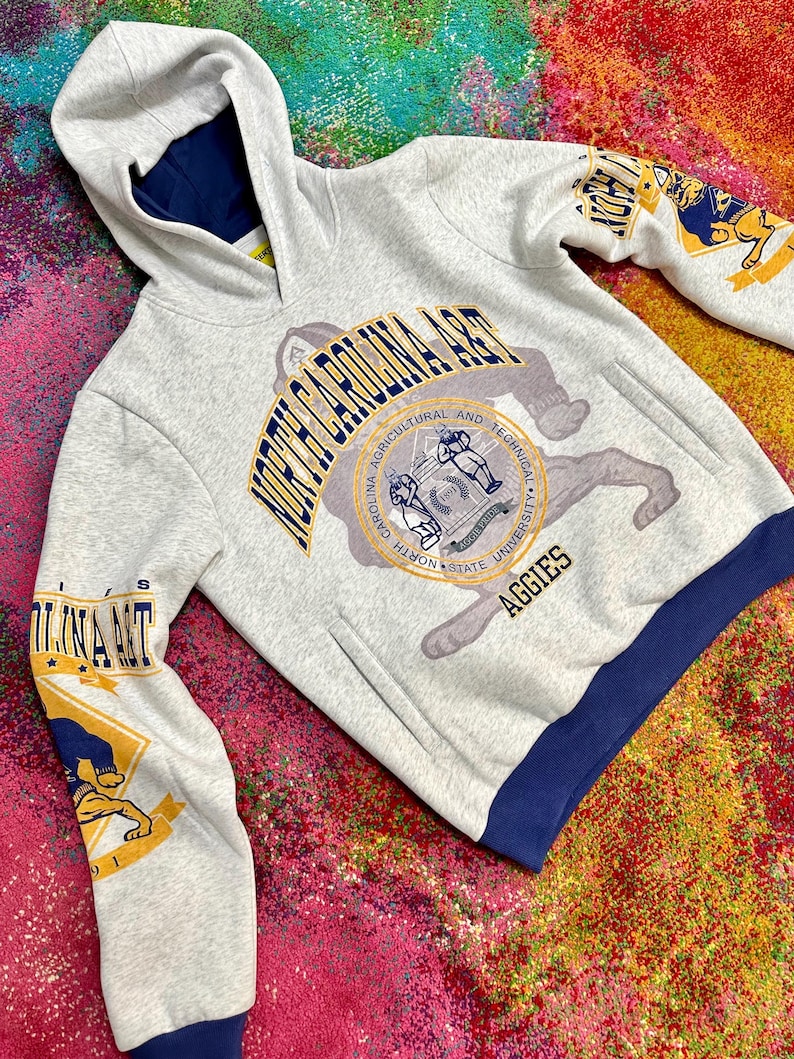 May include: A light gray hooded sweatshirt featuring North Carolina A&T branding. The front displays the university seal and the text North Carolina A&T Aggies. The sleeves feature graphic prints of a bulldog mascot, and the hem is dark blue.